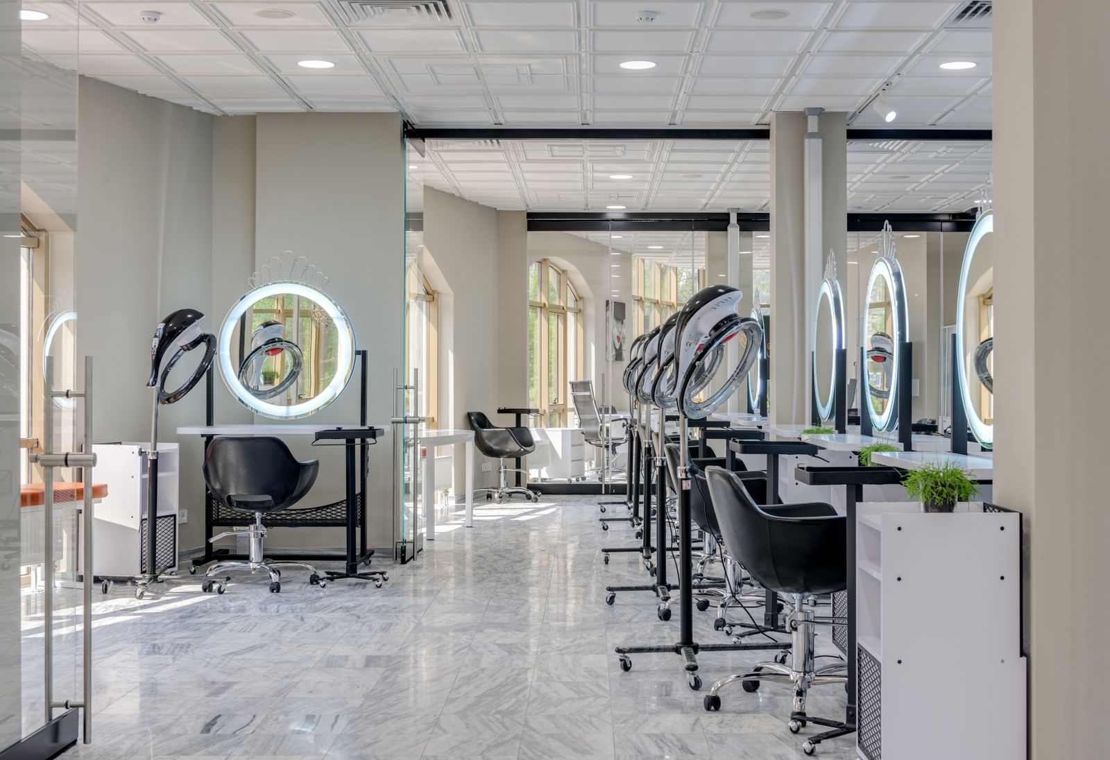 Bright, spacious salon with a pale marble-look floor, large arched windows at the back, a row of white styling stations each with a round ring-lit mirror, black swivel chairs with chrome bases and tall hood dryers on stands.