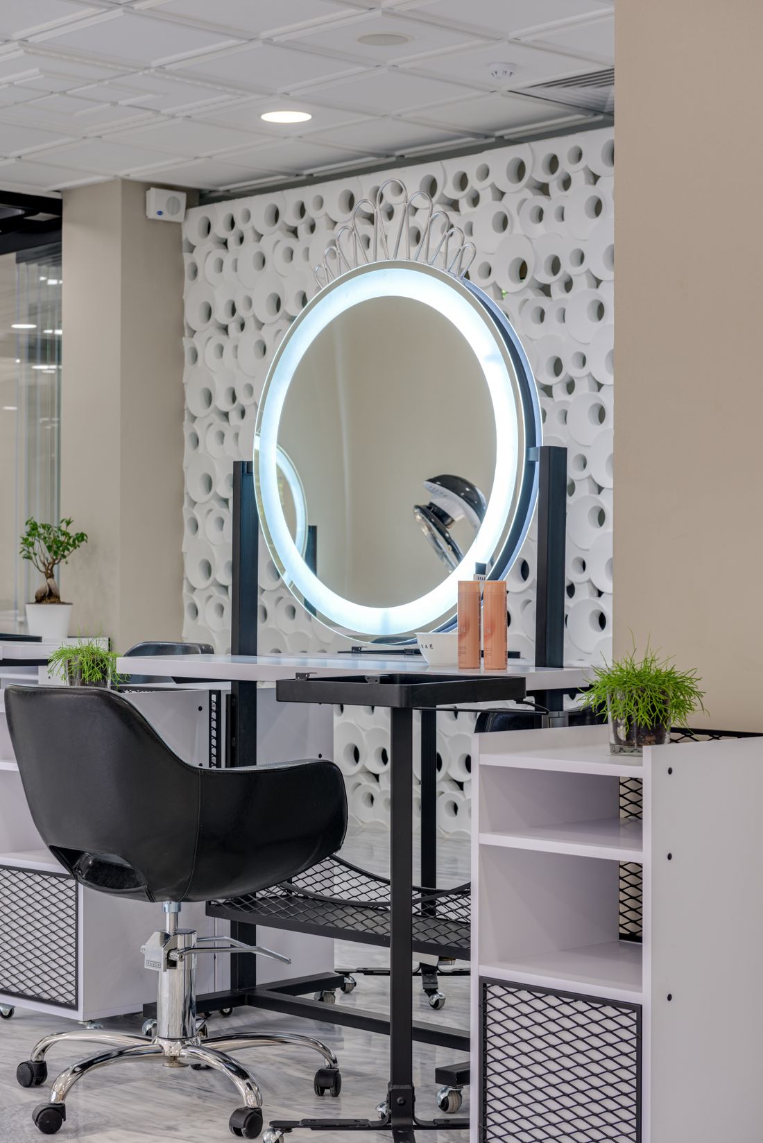 One styling station: a round LED ring-lit mirror with a small crown ornament on top, in front of a white wall of round cut-out patterns; a black styling chair at left, a black-and-white trolley with two unlabelled orange-brown bottles and a small white bowl, a potted bonsai, a green plant in a glass pot and a white open shelf unit at right.