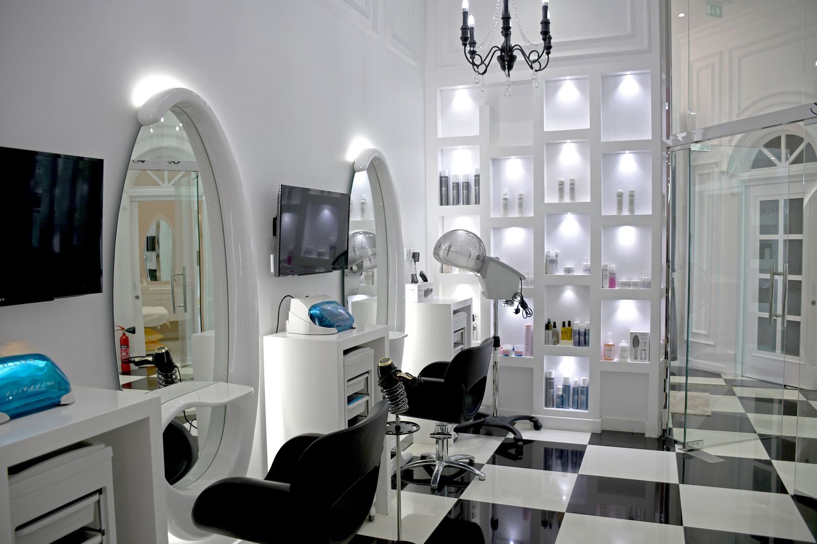 White salon with oval wall mirrors and wall-mounted screens, white treatment tables, black styling chairs, a hood dryer, a lit white shelving wall holding unlabelled product bottles, a black candle-style chandelier and glass doors to a second room, on a black-and-white checkerboard floor.